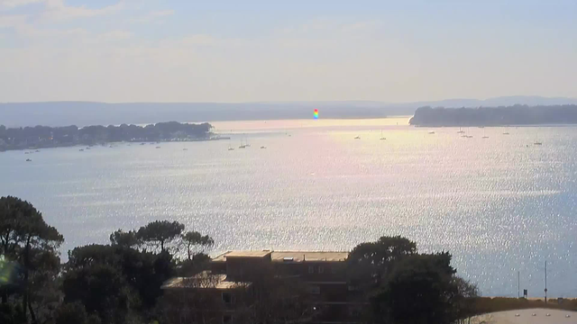 A scenic view of a calm body of water reflecting sunlight. In the foreground, there are some trees and a building near the water's edge. The middle ground shows several boats scattered across the water, while the background features distant hills and a shoreline. The sky is clear with a few clouds.