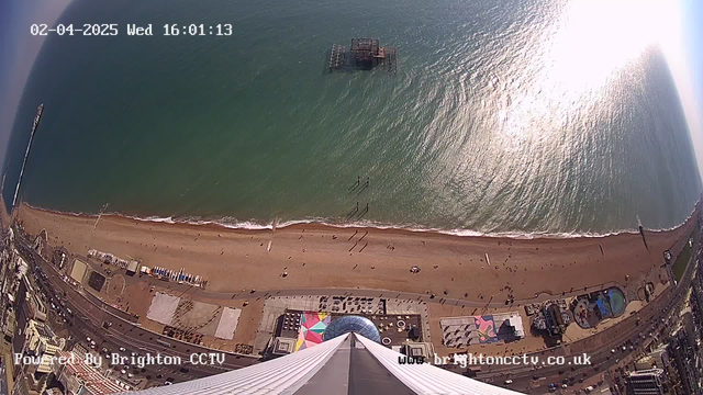 A high-angle view of a beach and seafront, with the ocean in the background reflecting sunlight. The beach is sandy with several people walking and relaxing along its length. There's a pier structure partially submerged in the water. Below the beach, various colorful tents and attractions are visible along the shorefront. A road runs parallel to the beach with parked cars and buildings lining the street, giving a vibrant seaside atmosphere.