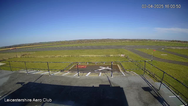 A clear blue sky stretches over a green landscape. In the foreground, there is a flat rooftop with a railing, and a helipad marked with white lines and a red section. To the right, there is an airstrip with a windsock indicating wind direction. The background features a vast area of grass, and in the far distance, rolling hills can be seen. The time displayed is 16:00 on April 2, 2025.