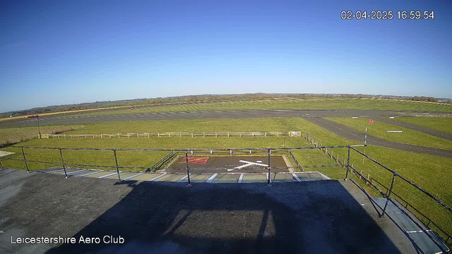 A clear blue sky stretches across the top of the image. Below, a wide open space features a runway with distinct markings. To the left, there is a grassy area enclosed by a white wooden fence. In the foreground, there is a view of a flat rooftop where the webcam is positioned, casting a shadow on the surface. The scene is bright, indicating sunny weather. In the distance, rolling hills are visible on the horizon.