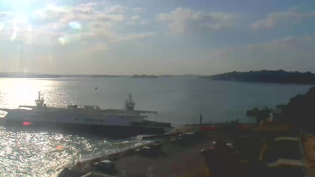 A sunny view of a calm body of water reflecting light. In the foreground, a large ferry boat is docked. There are several cars parked along the edge of the dock, and in the background, there are distant hills and a clear blue sky with a few clouds. The scene conveys a peaceful maritime atmosphere.
