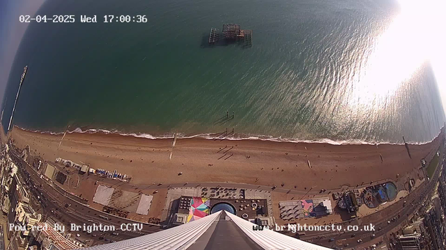 A high-angle view of a beach on a sunny day. The sandy shoreline stretches out below, with groups of people walking along the beach. Gentle waves lap at the shore, and a pier extends into the water. An old, partially submerged pier structure is visible in the sea. Nearby, colorful beach structures and tents can be seen along the promenade. The image captures a clear sky and reflects sunlight on the water's surface. The date and time are displayed in the corner of the image.