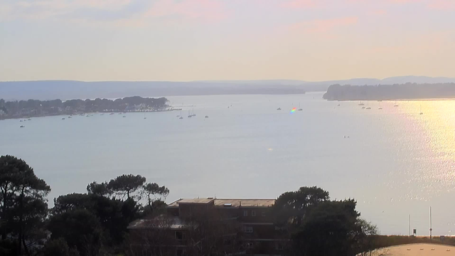 A serene view of a body of water reflecting the sunlight, with a soft gradient in the sky from pale blue to light pink. Small boats are scattered across the water, and a distant shoreline is lined with trees and buildings. The scene is tranquil, capturing a calm atmosphere with gentle waves and a few ripples near the shore.