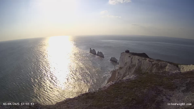 A panoramic view of the ocean under a hazy sky, with the sun reflecting off the water, creating a shimmering effect. In the foreground, grassy cliffs lead down to the sea. Two prominent rock formations rise from the water, and a building is visible at the top of the cliff on the right side. The image has a serene atmosphere with soft colors.
