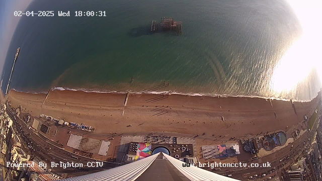 An aerial view of a sandy beach extending along the shoreline. There are people walking on the beach, and some are near the water's edge. To the right, there is a partially visible amusement area with colorful structures. In the distance, a pier extends into the water, and a structure is visible on the ocean's surface. The scene is illuminated by sunlight, creating reflections on the water. The date and time are displayed in the top left corner, and the bottom of the image includes a watermark for the camera source.