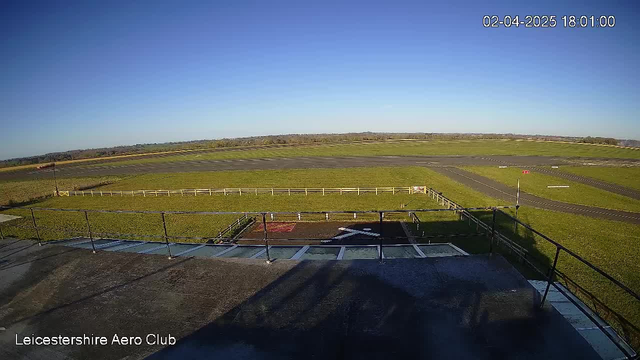 A clear blue sky stretches overhead, with no clouds visible. Below, a grassy field is seen featuring a runway. There are markings on the ground indicating takeoff and landing areas, along with a white cross shape in one section. In the distance, a flat landscape extends, showing patches of green grass, and some outlined areas possibly for parking or other airport activities. The scene is tranquil and open, indicating a wide expanse of land and sky.