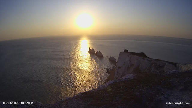 A sunset view over the ocean, with the sun glowing brightly above the horizon, casting a golden reflection on the water. In the foreground, there are rugged cliffs with some rocky formations jutting out into the sea. A small structure is visible on top of one of the cliffs, surrounded by the natural landscape. The sky transitions from blue to orange as the day ends.