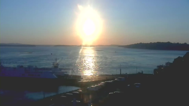 A scenic view of a body of water under a bright orange and white sun, setting in the horizon. The sunlight reflects on the water, creating a shimmering effect. In the foreground, there is a parking area with several cars visible, and a boat docked nearby. The background features a distant shoreline with trees and hills silhouetted against the sky. The overall scene conveys a calm and peaceful atmosphere.