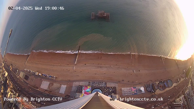 A high-angle view of a beach with a pier extending into the water. The sandy beach is lined with a variety of structures, including amusement rides and colorful tents. The ocean is visible, with gentle waves lapping at the shore. In the background, there is a faint outline of the pier structure, partially submerged, and the scene is bathed in soft evening light. The date and time are displayed in the top left corner.