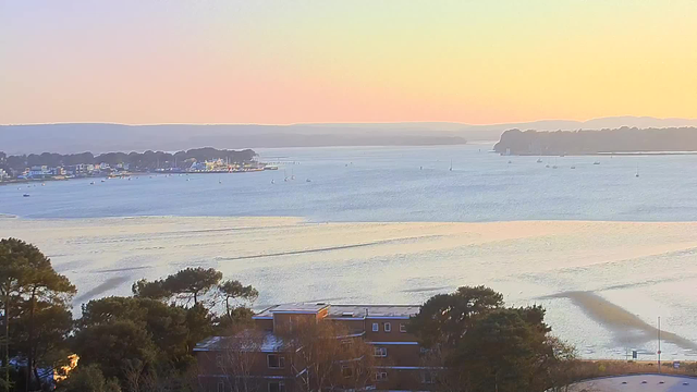 A serene coastal scene during sunrise, featuring calm waters reflecting soft pastel colors of orange, pink, and blue. In the foreground, there are tree tops and a low building. In the distance, small boats are scattered on the water, with a shoreline lined by houses. The background shows gentle hills and more greenery along the coast under a clear sky.