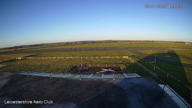 A clear blue sky is visible above an expansive grassy field, which is part of an airstrip. In the foreground, a tarmac area with a white cross marking and a small circular object can be seen. To the left, there is a fence bordering the field, and a windsock is positioned on a pole, indicating wind direction. The scene is peaceful, with no aircraft in sight. The image's timestamp shows it is early evening.