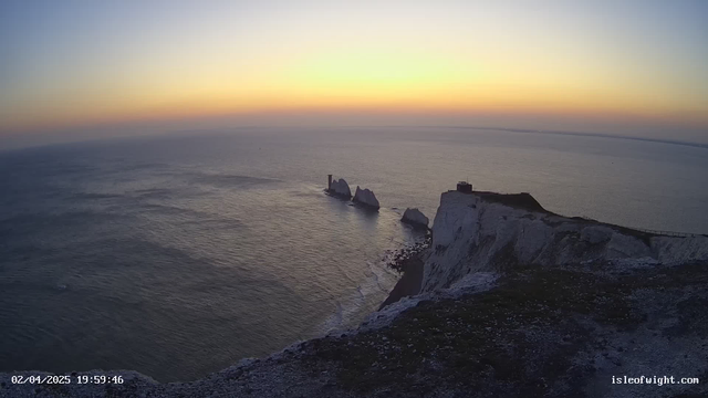 A scenic view of the coastline at sunset. The horizon features soft gradients of yellow, orange, and pale blue. In the foreground, there are chalk cliffs with some green vegetation. To the right, a small structure sits at the top of the cliffs. Below, jagged white rocks emerge from the sea, and gentle waves lap at the shore. The water appears calm, reflecting the sky's colors.