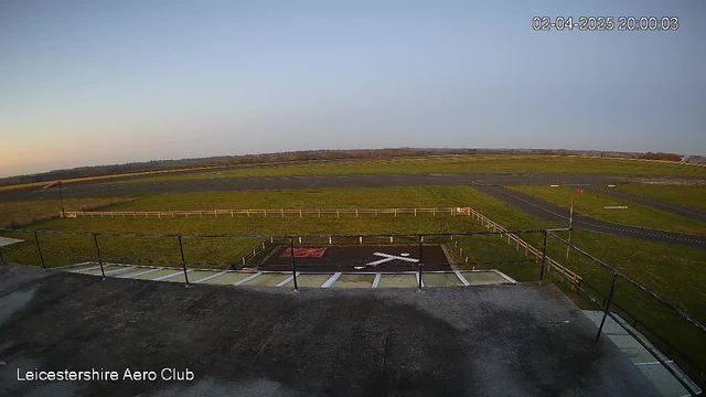 A wide-angle view of an airfield at sunset. The sky is clear with soft gradient colors transitioning from blue to a warm orange. The field is mostly green grass, bordered by a white wooden fence. A small section of the runway is visible, with a painted white "X" marking on the ground, indicating an area not in use. In the distance, there is a road and trees lining the horizon. The scene is tranquil, with no aircraft present. The image is timestamped at 8:00 PM on February 4, 2025.