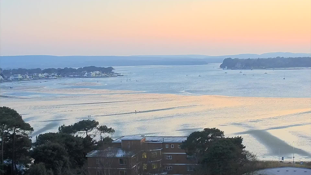 A serene landscape showing a river estuary at sunset. The sky transitions from light orange to soft blue, reflecting on the calm water. There are a few small boats scattered across the water, and silhouettes of trees line the shore. In the foreground, there is a building partially obscured by trees. The overall scene conveys a peaceful and tranquil atmosphere.