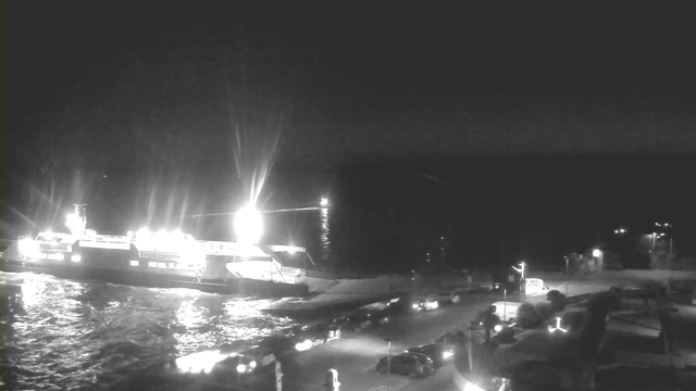 A dark coastal scene at night featuring a brightly lit ferry docked at a pier. The ferry is positioned to the left of the image, surrounded by shimmering water reflecting its lights. In the foreground, several vehicles are parked along a road next to the pier, with occasional streetlights illuminating the area. The background shows a dark shoreline, with a faint outline of land and a small distant light on the water.