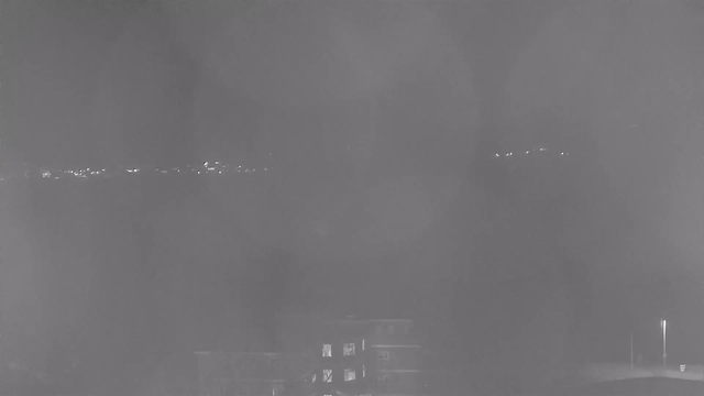 A low-visibility scene captured in black and white, showing faint outlines of distant lights on the horizon, possibly representing a city. In the foreground, there is a dark building with illuminated windows. The overall atmosphere is hazy, with indistinct shapes and varying light intensities contributing to a soft, blurred effect.