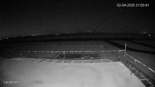 A nighttime view from a webcam positioned on a rooftop at the Leicestershire Aero Club. The image shows a dark sky with faint light spots in the distance, suggesting distant buildings or stars. In the foreground, there is a flat surface with railing, and a section that appears to have a crosshatch marking, possibly indicating a designated area. The overall scene is dimly lit with minimal detail due to the low light conditions. The time and date in the corner reads "02-04-2025 21:00:41."