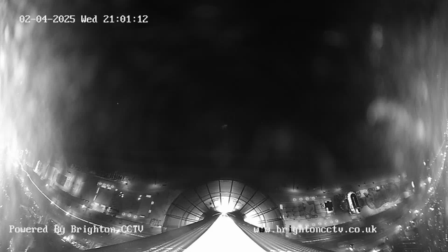 A night view from a webcam looking down at a cityscape. The image is predominantly dark with some illuminated areas. In the foreground, there is a bright pathway leading downward, resembling a ramp or tunnel. The background shows blurred outlines of buildings and streets, with faint lights indicating activity below. The time and date are displayed in the upper left corner, indicating it is April 2, 2025, at 21:01:12. The bottom left corner includes a watermark with the text "Powered By Brighton CCTV" and a website link in the bottom right corner.