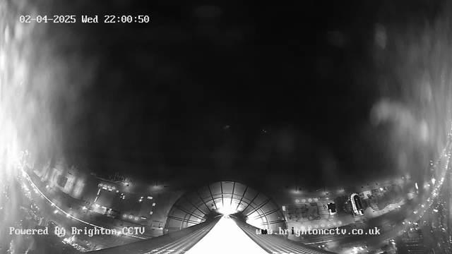 A black and white webcam image showing a view from a tall building or structure. The scene is mostly dark with a faint glowing area at the bottom, suggesting a lit pathway or street. There are faint outlines of buildings and possibly vehicles along the edge of the image, indicating an urban environment. The timestamp in the upper left corner reads February 4, 2025, at 10:00:50 PM. There is text in the lower left corner indicating the image is powered by Brighton CCTV.