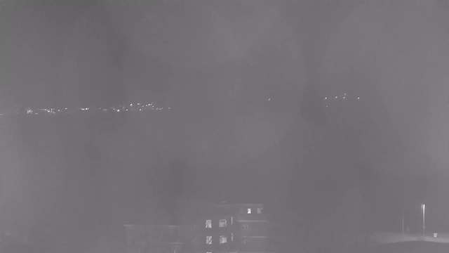 A nighttime webcam view shows a dark scene with faint lights from buildings in the distance along the horizon. In the foreground, there is a structure with a lit window, and a streetlight is visible nearby. The overall image has a low contrast with a grayish tone, creating a somewhat foggy or hazy appearance.