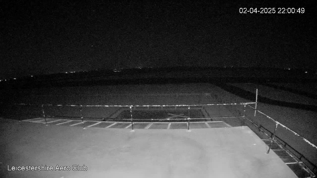 A dark outdoor scene from a webcam at Leicester Aero Club, showing a flat surface in the foreground with a railing along the edge. The background is mostly black with subtle hints of distant lights, likely representing a horizon or far-off landscape, indicating it is nighttime. The time stamp in the corner shows the date and time as April 2, 2025, at 10:00 PM.