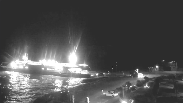 A ferry is illuminated at night, docked at a port. The water is reflecting light from the ferry and nearby streetlights. There are vehicles parked near the edge of the water, with a few more approaching the area. The scene is predominantly dark with bright spots of light, creating a contrast against the night sky.