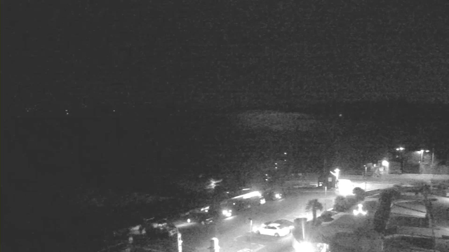 A dimly lit coastal scene captured at night. The image shows some lights from vehicles parked along a road, with a few streetlights illuminating parts of the area. In the distance, there are faint, scattered lights possibly indicating buildings or boats. The water appears dark, with no visible details due to the low visibility. The overall atmosphere is quiet and serene, with a few shadows obscuring the surrounding features.