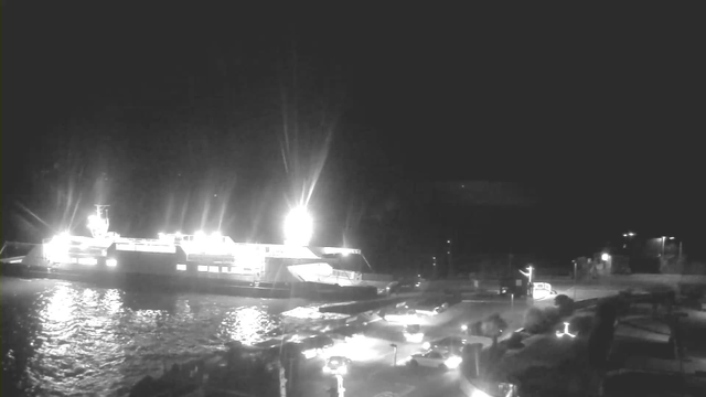 A brightly lit ferry is docked at a port at night, with its lights reflecting on the water. There are several vehicles parked near the edge of the dock, and additional lights from the surrounding area are visible, creating a dimly illuminated scene. The sky is dark, enhancing the contrast between the light from the ferry and the shadows of the surroundings.