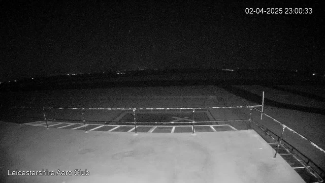 A dark scene from a webcam at Leicestershire Aero Club, showing a flat rooftop surface with a railing along the edge. In the background, there are faint lights visible in the distance, likely from surrounding areas. The image is timestamped with the date and time: April 2, 2025, at 11:00 PM.