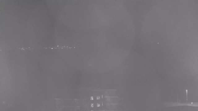 A dimly lit scene captured from a webcam at night. The foreground shows an indistinct building with some lit windows, while the background features a blurred skyline with small pinpricks of light representing distant structures or reflections. The overall image has a soft focus, predominantly in shades of gray.