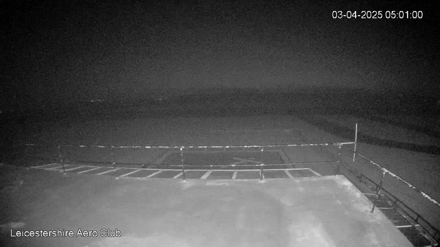 The image shows a dimly lit scene from a webcam at the Leicestershire Aero Club, timestamped 03-Apr-2025 at 05:01. The foreground features a flat surface with markings and a low barrier, while the background is dark with minimal visibility, suggesting it may be nighttime or early dawn. There are faint outlines of the horizon or distant features.