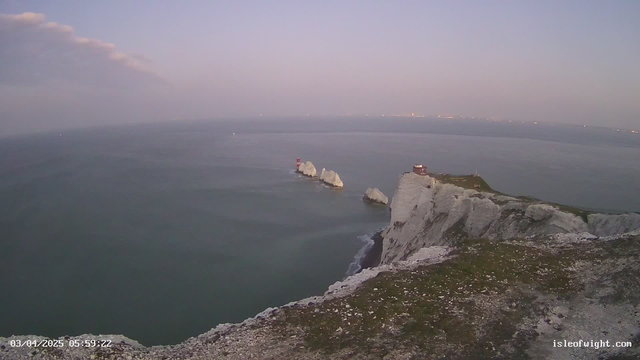 A coastal scene at dawn, featuring white chalk cliffs rising from the ocean. In the foreground, grassy land leads to the edge of the cliff, with rocky outcrops emerging from the water. A small red lighthouse is positioned on a rocky formation, while several white rocks are clustered nearby. The sea is calm, with a muted, blue-gray color reflecting the morning sky, which transitions from light to darker shades. Farther in the distance, faint lights from a town or city twinkle along the horizon.