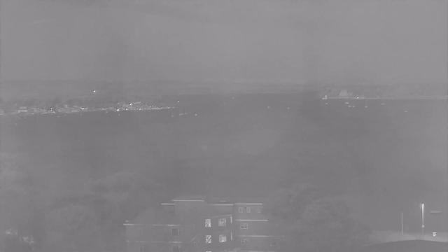 A grayscale image showing a distant view of a body of water. In the foreground, there is a low-rise building with multiple windows, partially obscured by trees. The water appears calm with a few small boats visible in the distance. The horizon line is faint, suggesting distant land or hills. Overall, the scene has a foggy or hazy quality with soft contrasts.