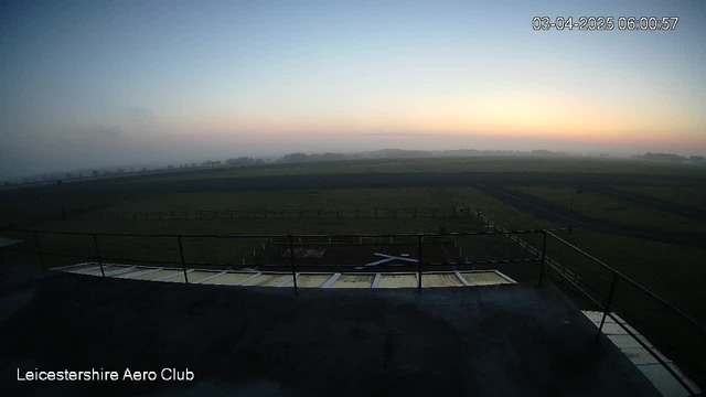 A panoramic view of a foggy landscape at dawn. The sky is a gradient of soft pastel colors, transitioning from pale blue at the top to warm hues of orange and yellow near the horizon. A wide, empty grassy field stretches across the scene, bordered by distant tree lines on the horizon. A large, unmarked runway is visible, with outlines of trees and fields beyond it. The foreground features a low railing along the edge of a platform, suggesting the view is from a higher elevation. A timestamp indicates the image was taken early in the morning.