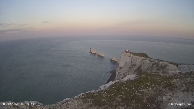 A coastal scene shows a rocky shoreline with steep cliffs on the right side. The cliffs are predominantly white and partially covered with grass. In the distance, several small rock formations emerge from the water, with a red lighthouse on one of them. The sky is light blue with soft pink hues near the horizon, indicating either dawn or dusk. The calm sea stretches out toward the horizon, reflecting the colors of the sky.