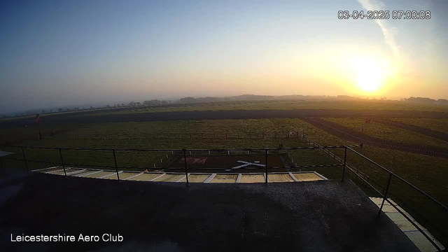 A sunrise scene over a green airport field, with a clear sky transitioning from blue to orange and yellow near the horizon. The foreground features a railing and a concrete surface, while the background shows a landing strip and an open expanse of grass. A flag is visible on the left side, and the distant landscape includes low hills and trees. The image timestamp indicates the date and time.