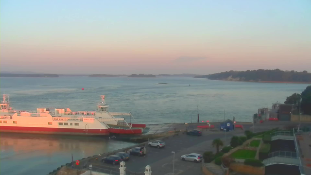 A wide view of a harbor during sunset, featuring a large white and red ferry docked at the edge of the water. The calm sea reflects soft hues of blue and pink from the sky. Several vehicles are parked nearby, and a small building can be seen to the right, along with some green landscaping. In the distance, faint outlines of land can be observed against the horizon.