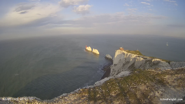 A panoramic view of the coastline featuring white chalk cliffs and the ocean. In the foreground, there are grassy areas and rocky outcrops. To the right, a lighthouse structure is visible, along with a small building atop the cliff. In the distance, a sailboat is seen on the water, and the sky is partly cloudy with patches of blue. The scene conveys a serene coastal landscape.