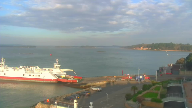 A ferry is docked at a terminal near the water. The scene shows calm waters with a few boats visible and a rocky shoreline in the distance. The sky is partly cloudy, creating a soft light over the area. There are cars parked near the ferry terminal, and some greenery is present along the waterfront.