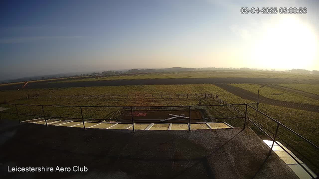 A wide view of an airport landscape under a clear sky at sunrise. In the foreground, there is a railing and steps leading down from a structure. The ground is mostly green grass, with a small section that appears to be a runway or landing area marked with a white X shape. In the background, a field with distant trees is visible, and the sky is lightening as the sun rises.