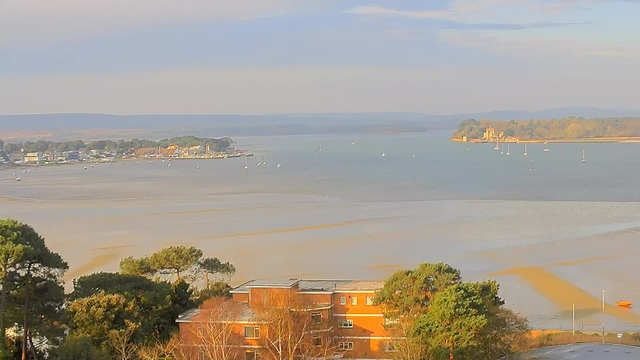 A tranquil waterfront view featuring calm waters extending into the distance, dotted with small boats. In the foreground, there is a low building surrounded by trees, and further back, a town with a mix of residential and commercial structures. Across the water, hills can be seen in the background under a light cloud cover. The scene is illuminated by soft natural light, suggesting it is either early morning or late afternoon.
