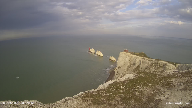 A coastal scene featuring steep white cliffs on the right, with the ocean stretching out to the horizon. In the middle of the image, two large white rock formations are visible in the water, one with a small red lighthouse on top. The sky above is mostly cloudy, with patches of blue peeking through. The foreground shows a grassy area with scattered pebbles, and the water appears calm with subtle waves.