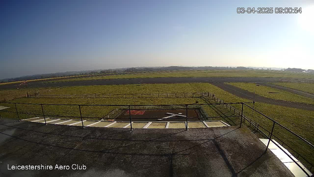 A wide view of a grassy airfield under a clear blue sky, with a visible tarmac runway and taxiway. In the foreground, there is a balcony railing and a helipad marked with a white cross on a dark surface. The airfield is bordered by a fence, and distant hills can be seen in the background. The scene conveys a peaceful, sunny day at the Leicestershire Aero Club.