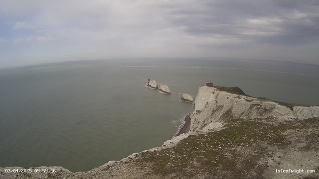 A coastal landscape featuring white cliffs meeting the sea under a cloudy sky. In the foreground, grassy terrain slopes down to a rocky shoreline. Two white rock formations rise from the water, with a red and white lighthouse positioned on one of them. The water appears calm and blue-green, with a slight wave pattern. In the distance, the horizon blends into the sky, creating a serene and natural seascape.