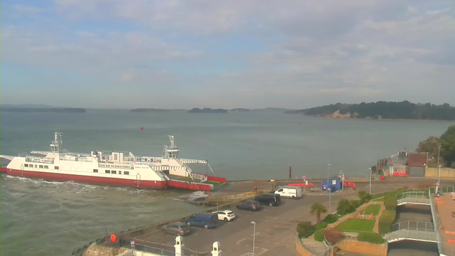 A ferry with a white and red hull is docked at a terminal, surrounded by calm waters under a partly cloudy sky. The terminal has a parking lot filled with cars, and there are landscaped areas with grass and palm trees nearby. In the background, small islands can be seen on the horizon.