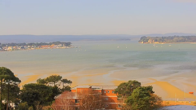 A panoramic view of a coastal scene featuring a calm sea with small boats scattered across the water. The background includes distant hills under a clear sky. In the foreground, there are trees and a red brick building, with sandy beach areas visible along the shore. The landscape is bright and tranquil, conveying a peaceful atmosphere.
