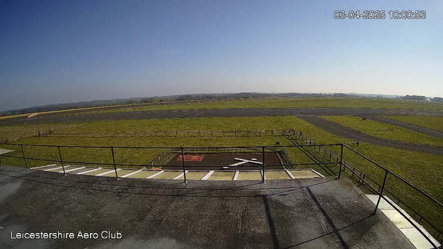 A clear blue sky is seen above a grassy field and an open area with a runway. In the foreground, a flat rooftop with a railing is visible. Near the center of the image, there is a large white X marking on a green surface, possibly indicating a landing area. The background features a horizon with a slight rise in the terrain, and a few distant structures can be seen. The timestamp at the top right corner indicates the image was captured on April 3, 2025, at 10:00:53.