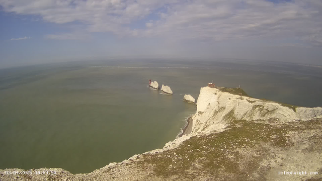A coastal view featuring white chalk cliffs extending into the sea. The water is calm and appears greenish, with some small waves. In the distance, there are two rocky formations with a red lighthouse on one of them. The sky is partly cloudy with light blue visible, and the foreground shows some grassy ground leading to the cliff's edge.