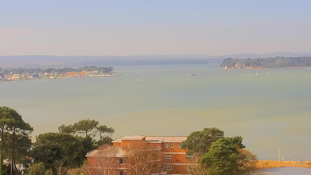 A scenic view of a calm body of water, with a shoreline lined by trees and buildings on the left. Various small boats dot the water, and in the distance, there are hills and a more distant landmass. The sky is clear with soft colors, suggesting a tranquil atmosphere. A few structures are visible in the foreground, including a brick building with multiple windows.
