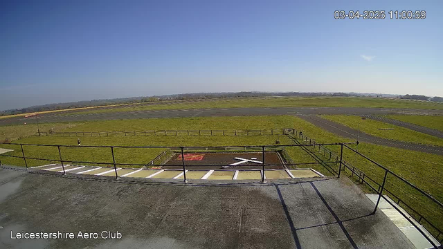 A wide-angle view from a webcam showing a grassy landscape with a runway. In the foreground, there is a flat roof with a railing. Below the roof, a red and white marking suggests a landing area with an 'X' shape made of white materials. The background features a clear blue sky and distant hills, with an expanse of green grass and a paved runway stretching into the distance. A fence marks the boundary of the area, and the time and date are displayed at the top right corner.